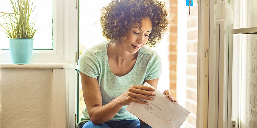 a woman returns home from the shops and finds some letters on the mat . She bends down to open them . coming home to bills